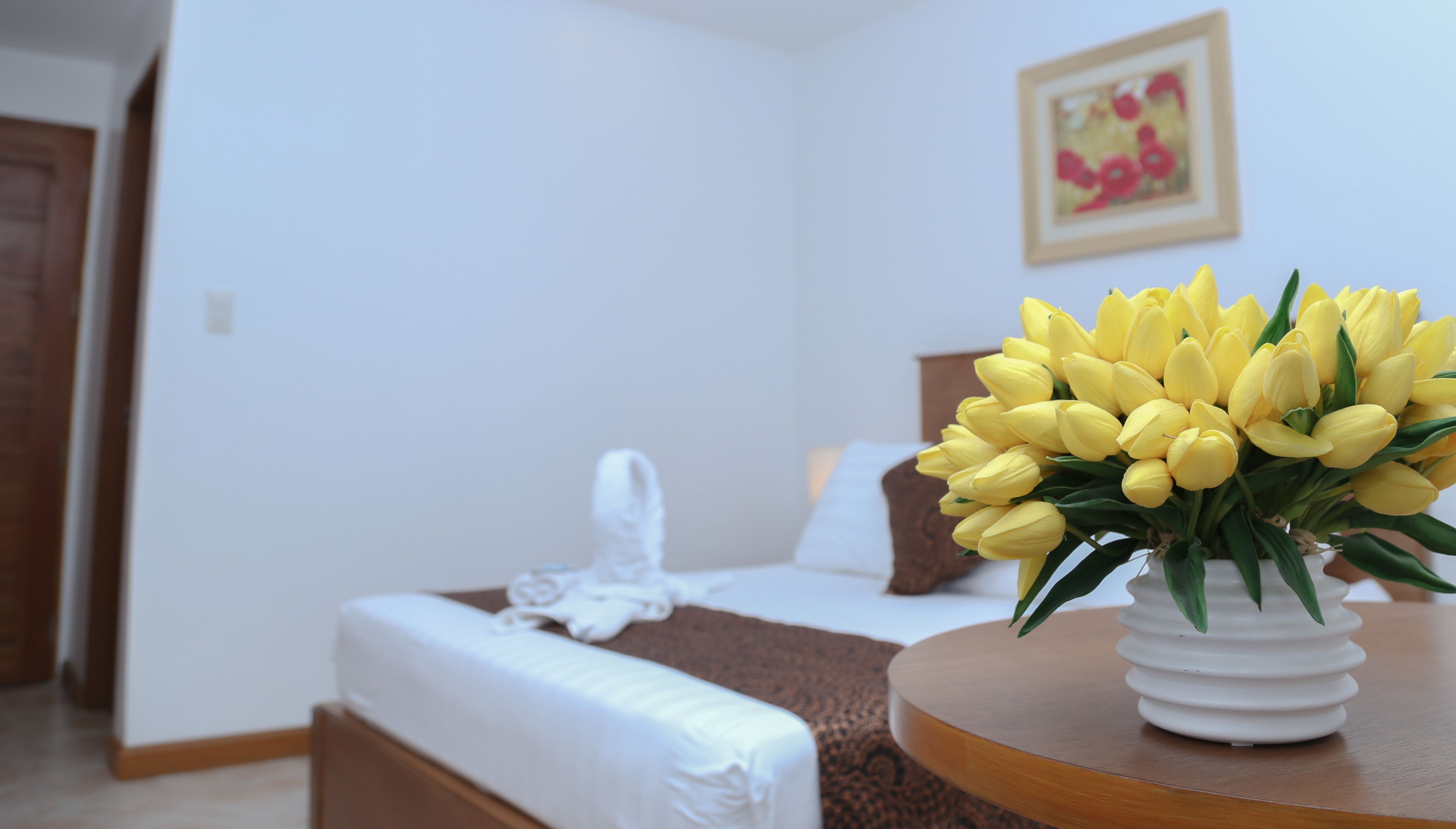 Una habitación de hotel con una cama, una silla blanca, una mesa blanca y un jarrón con flores amarillas.