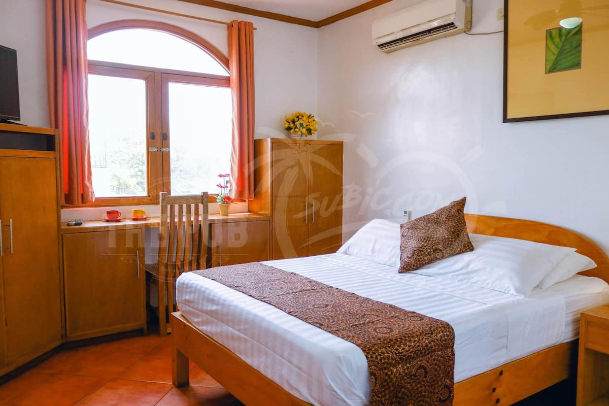 A bedroom with a bed, a window with curtains, a wooden wardrobe, a small table with a vase of flowers, and an air conditioning unit on the wall.