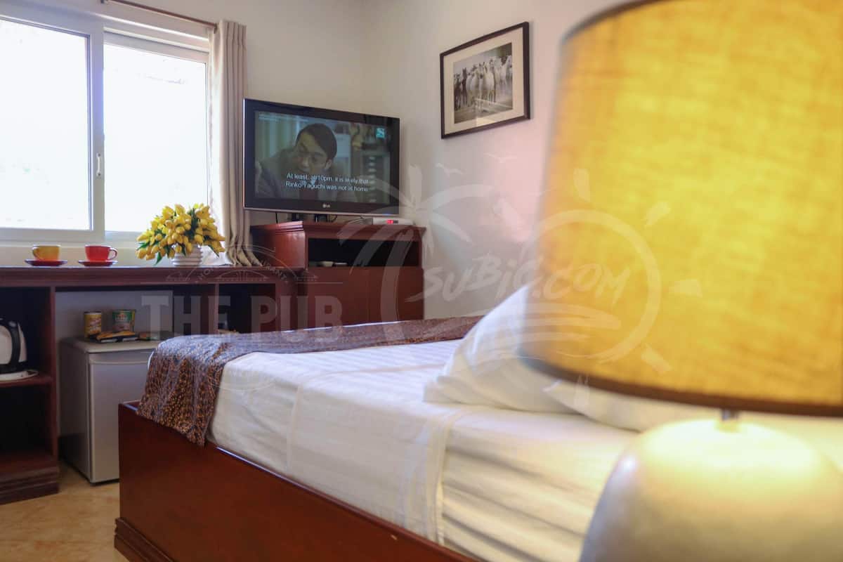 A hotel room with a bed, a television, a desk with a computer, a window, and a lamp.
