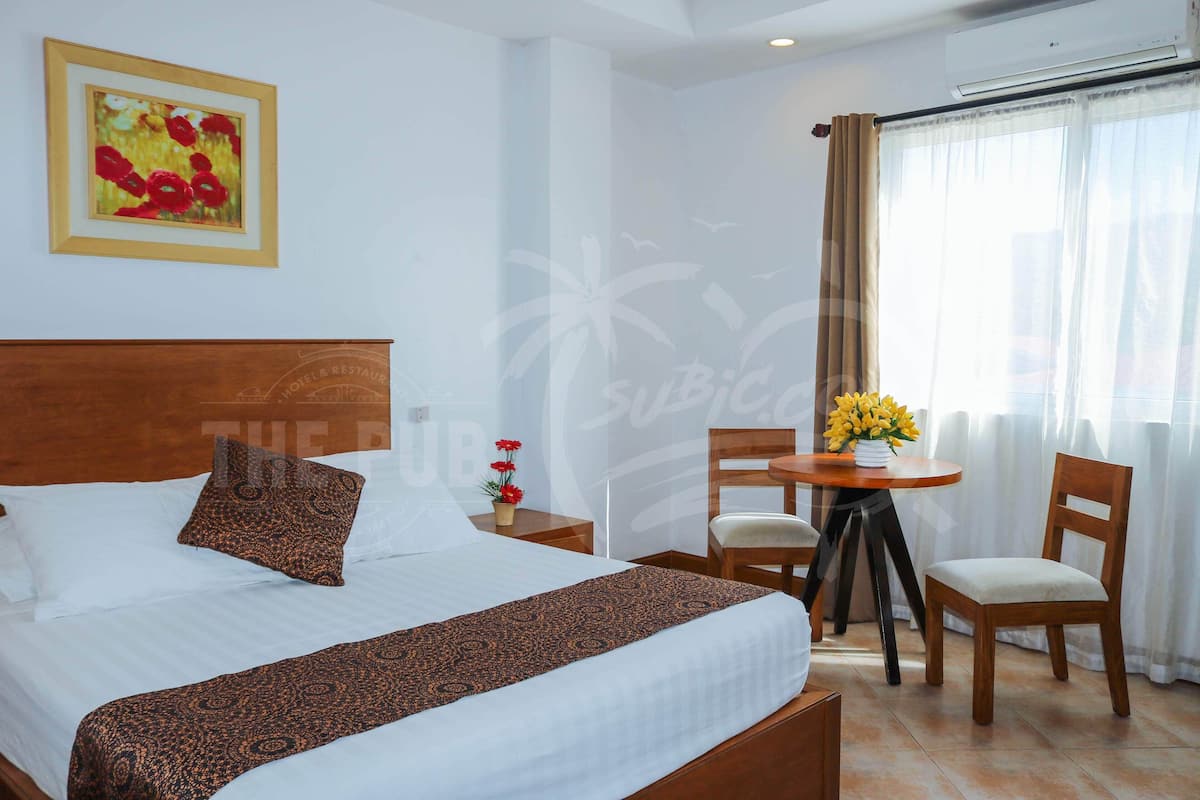 A hotel room with a bed, a small table with chairs, a vase of flowers, and a framed picture on the wall.