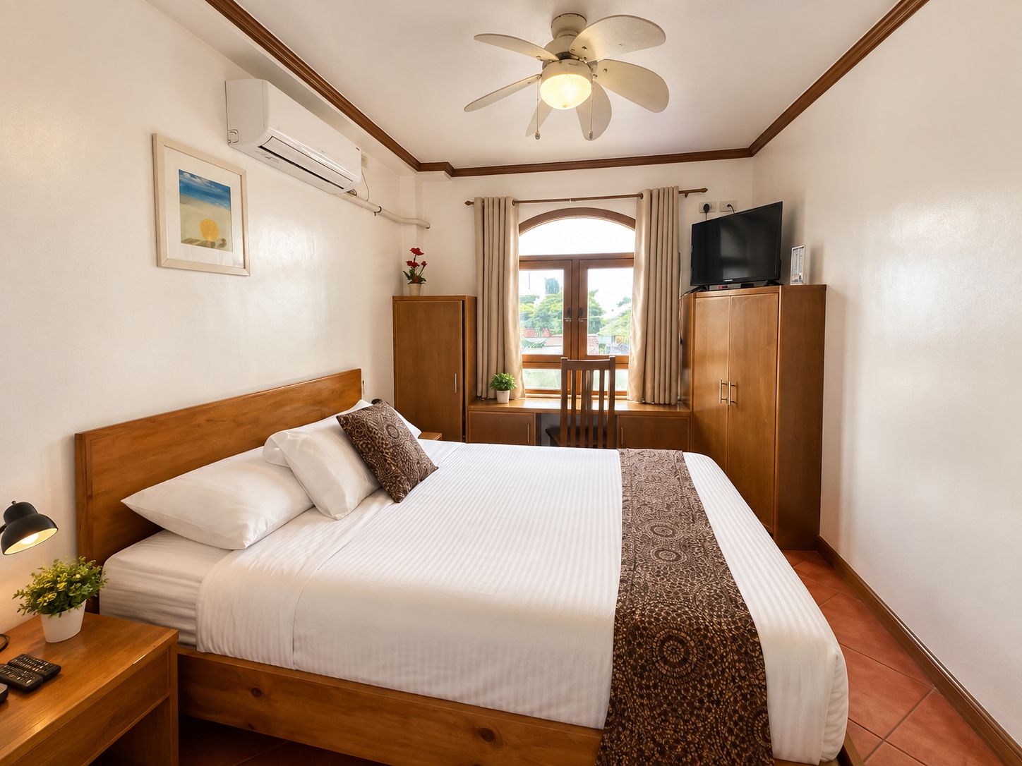 Una habitación de hotel con una cama, un escritorio con una lámpara, un televisor sobre un mueble y un ventilador de techo.