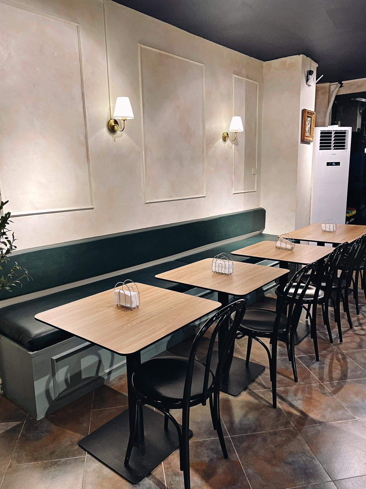 L’intérieur d’un restaurant avec des tables en bois et des chaises noires, un banc vert et des lampes fixées au mur.