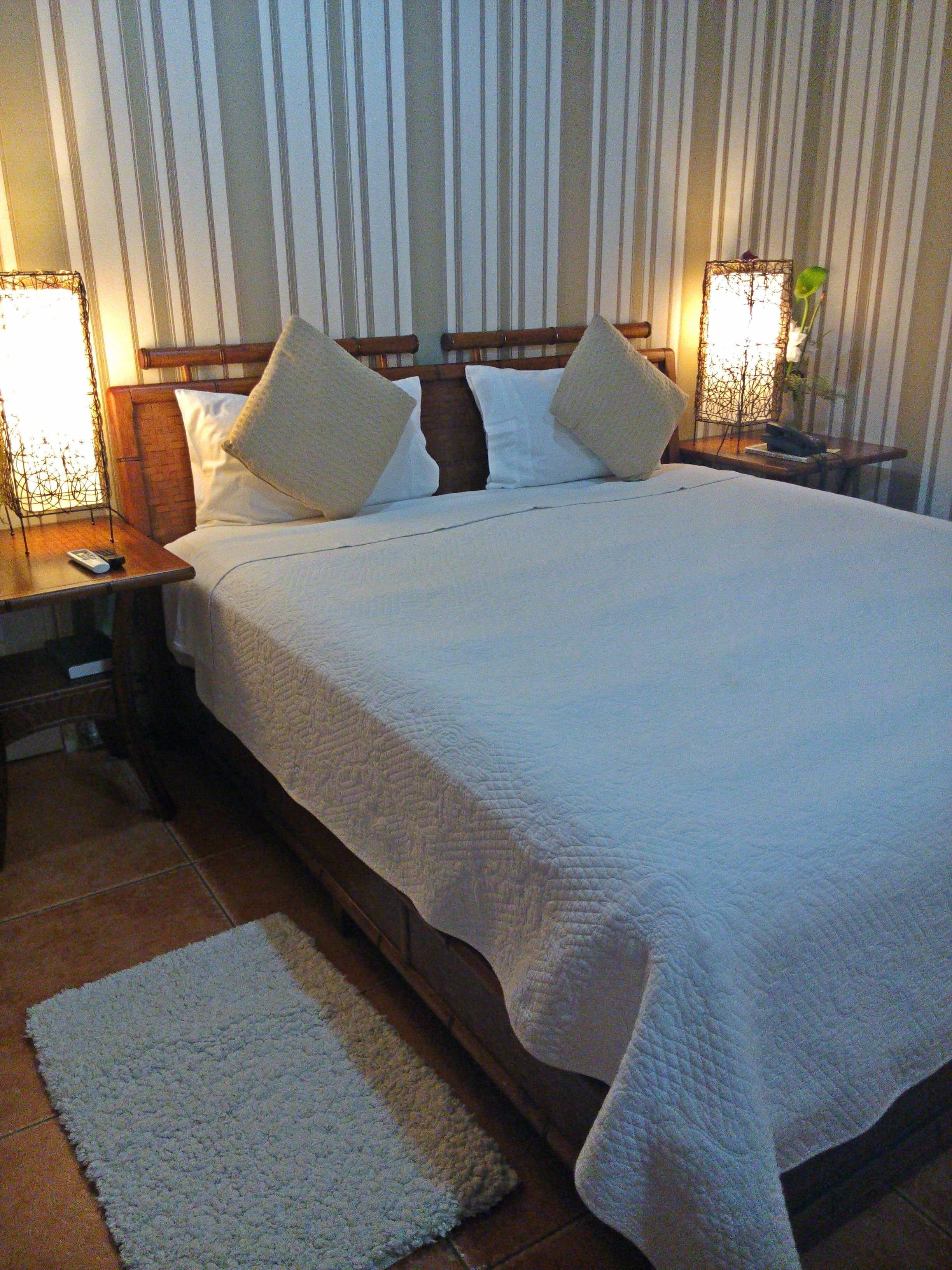 A neatly made bed with white bedding and two pillows, a wooden headboard, a bedside table with a lamp, and a small rug on the floor.
