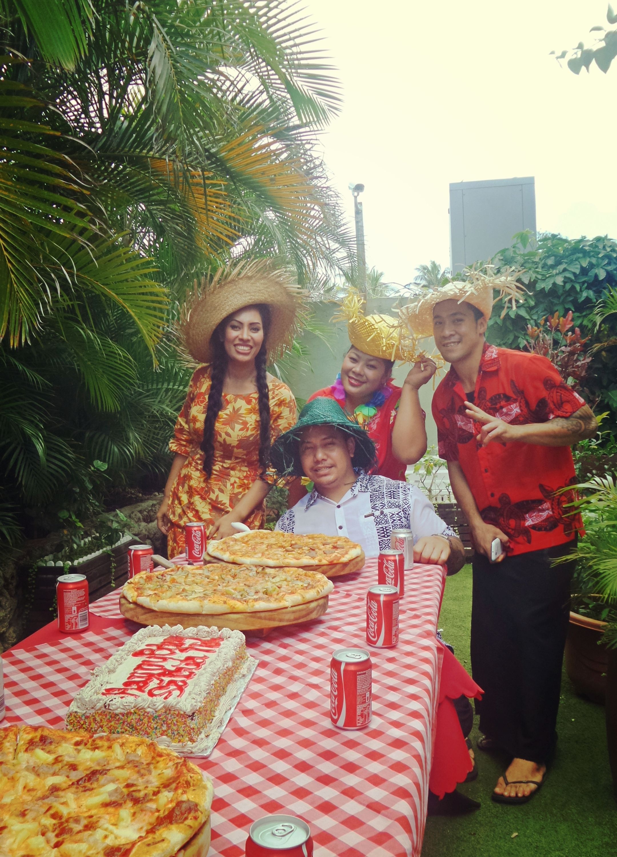 Um grupo de pessoas em um evento ao ar livre, com uma mesa posta de pizzas e latas de Coca-Cola.