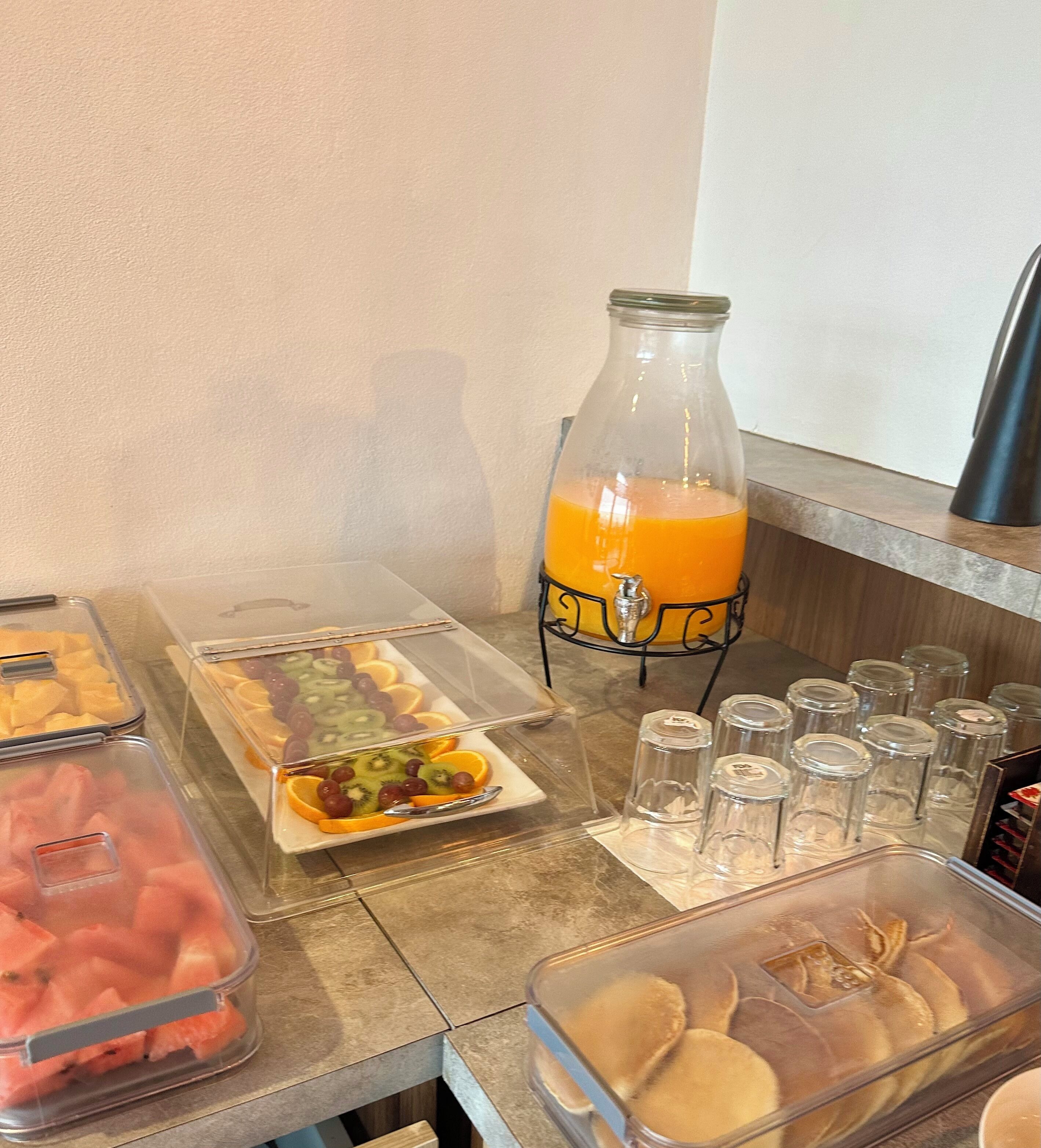 A breakfast buffet with various fruits, a glass of orange juice, and several condiment containers.
