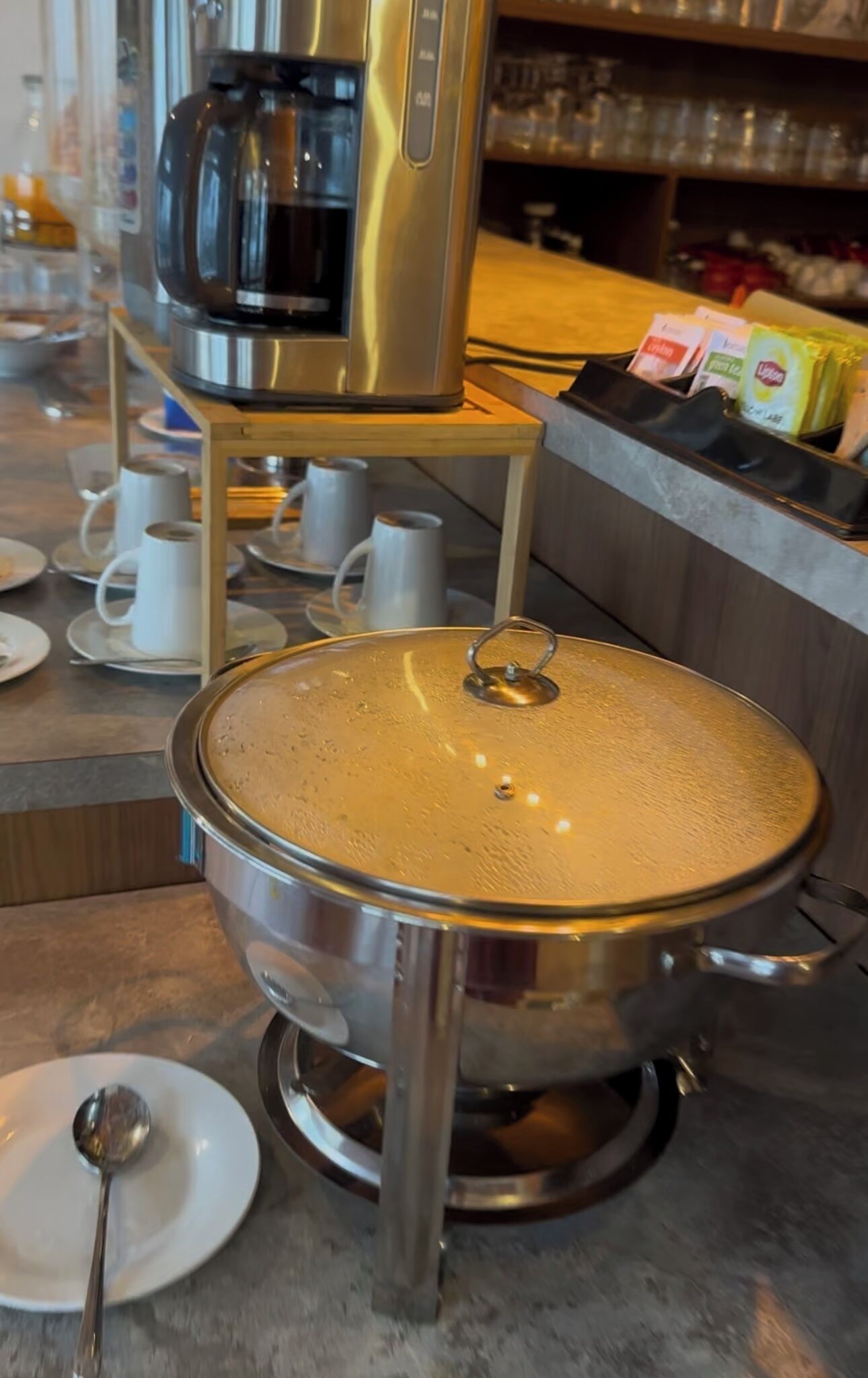 A stainless steel pot with a lid on a stand, a coffee machine, and a tray with disposable cups and a spoon.