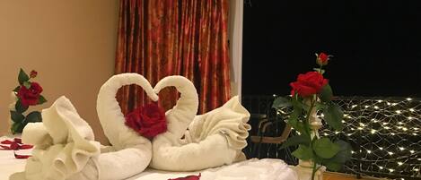 A bed with white linens, a heart-shaped towel arrangement, red roses, and a balcony with string lights.