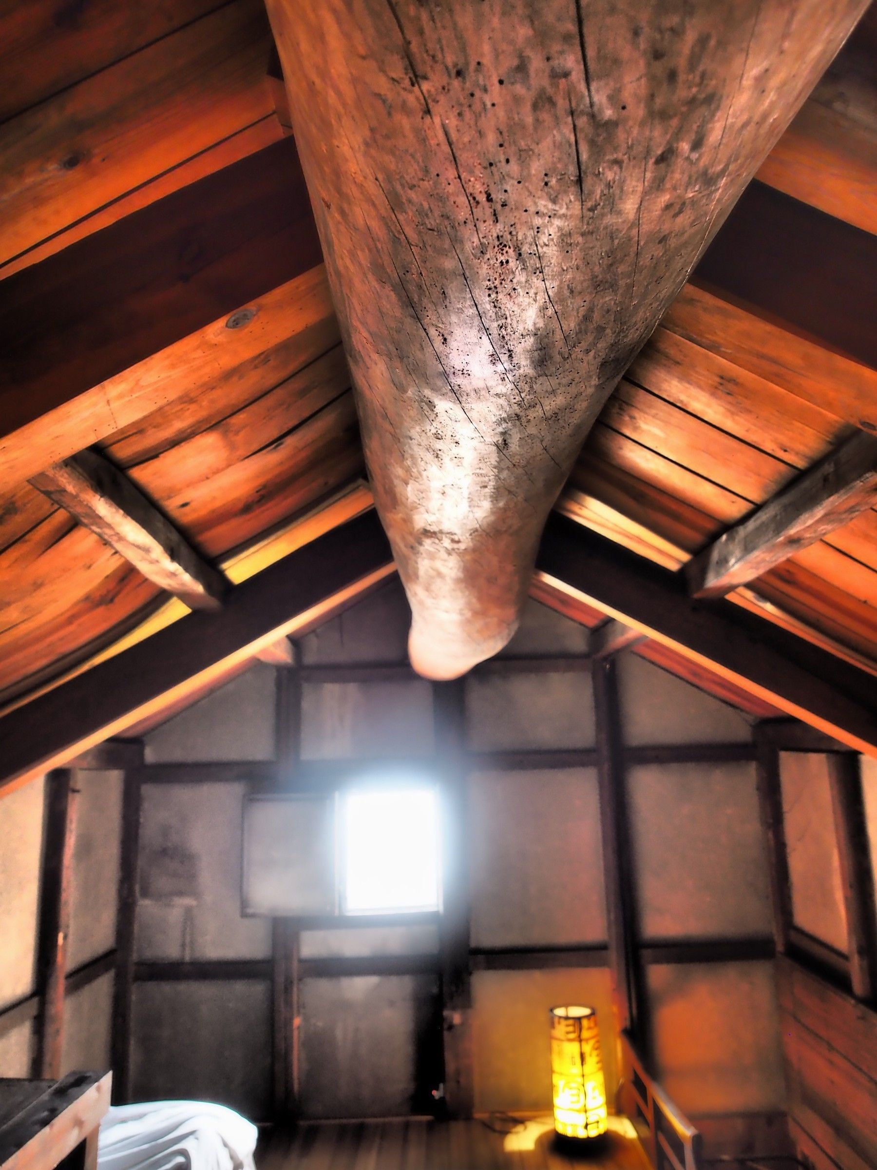 A rustic attic room with exposed wooden beams, a slanted ceiling, and a single window allowing natural light.