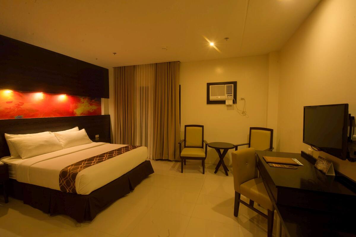 Habitación de hotel con cama, escritorio, dos sillas, televisión y aire acondicionado instalado en la pared.