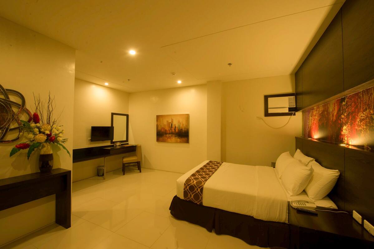 Una habitación de hotel con cama, mesita de noche, televisión, escritorio, silla y obras de arte en las paredes.