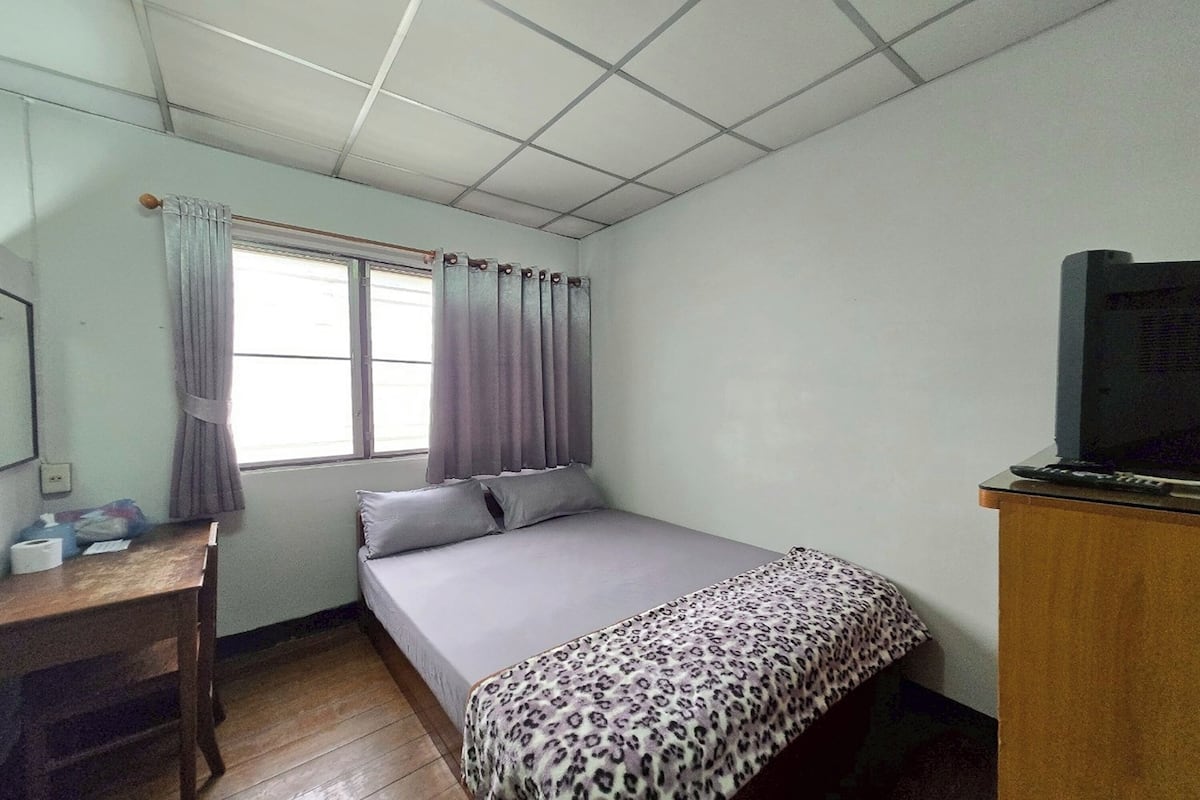 Une petite chambre avec un lit simple, un bureau en bois, un téléviseur posé sur une commode en bois et une fenêtre avec des rideaux.