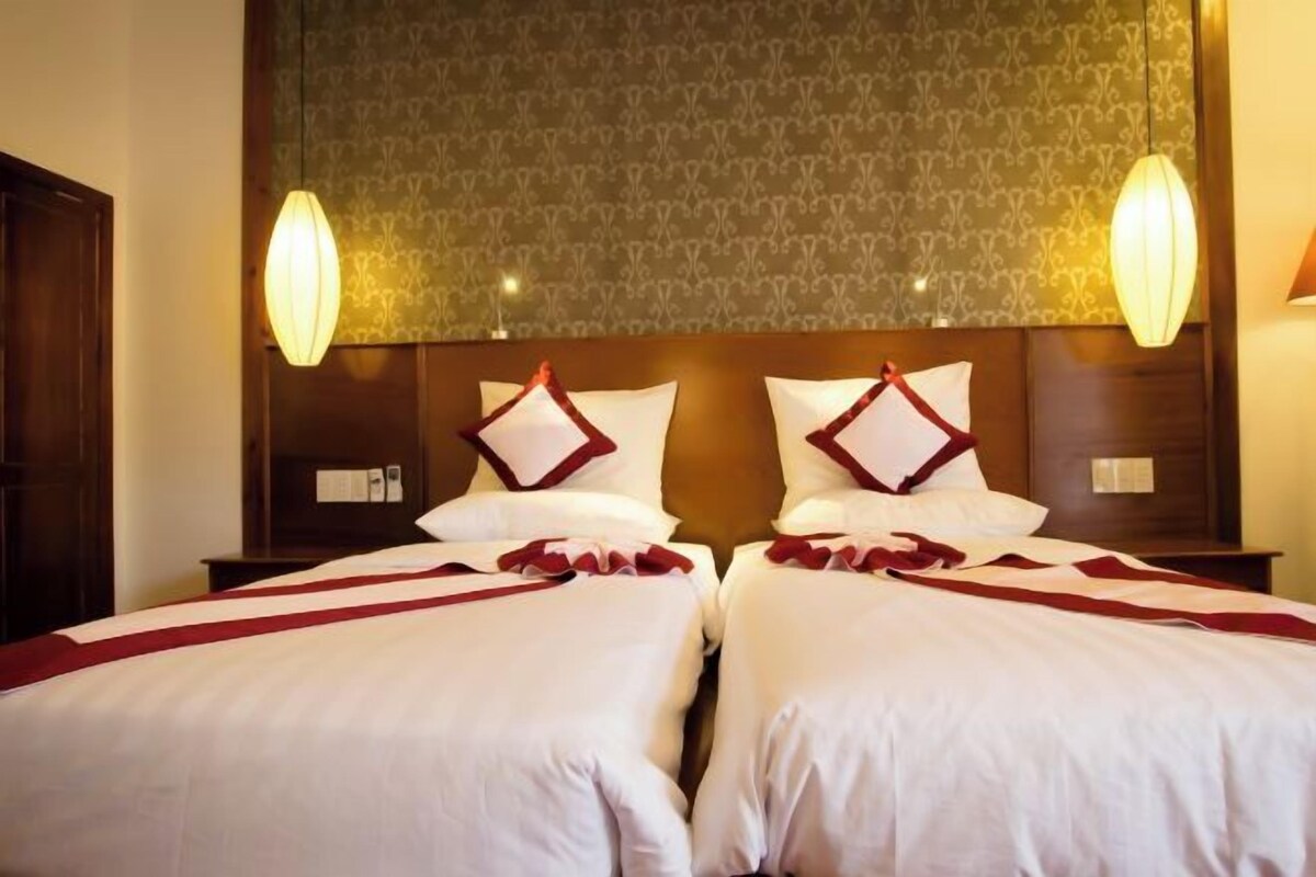 Une chambre d’hôtel avec deux lits, chacun agrémenté de draps blancs et de touches de couleur rouge, d’un papier peint à motifs et de deux lampes fixées au mur.