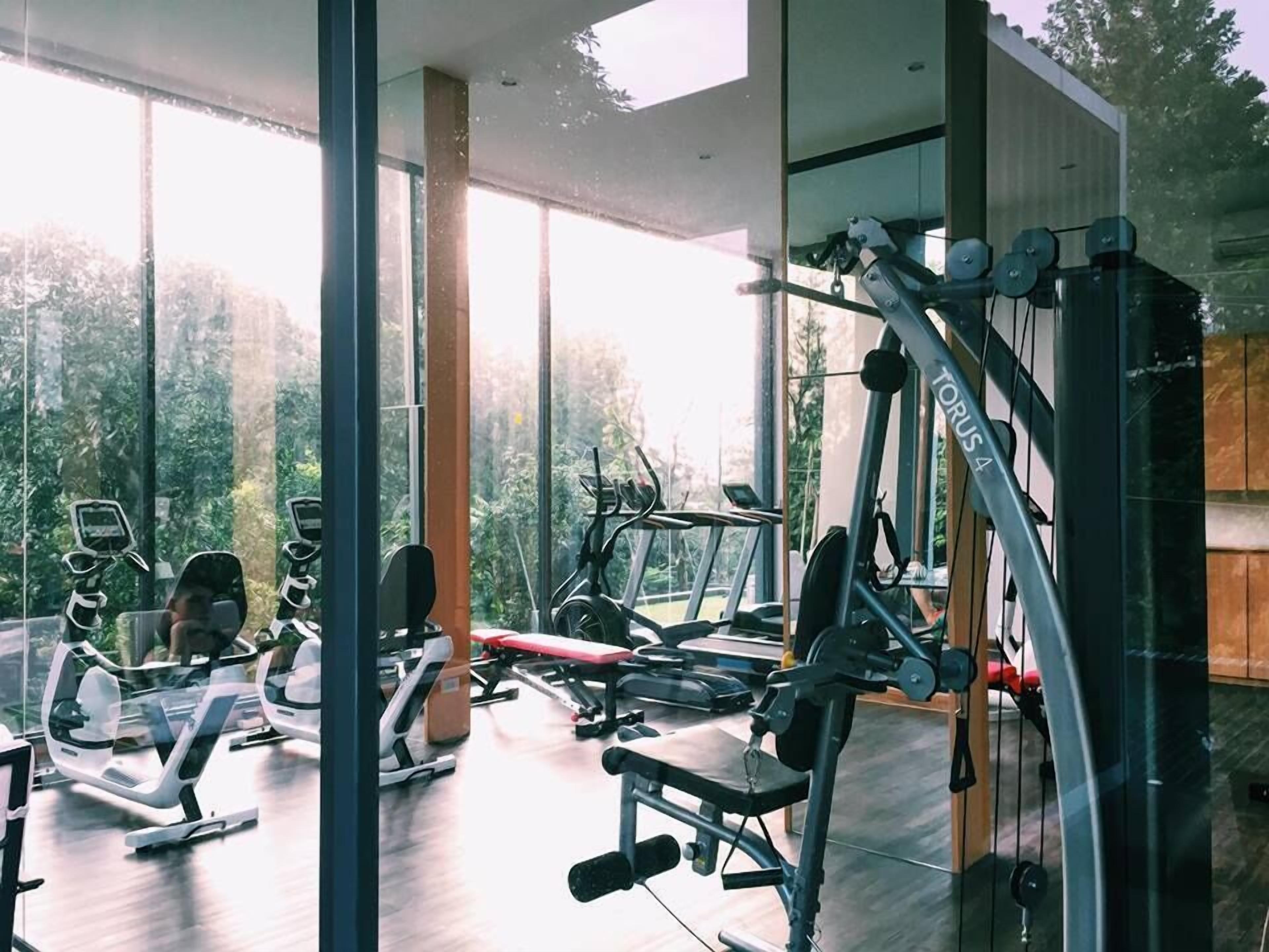 Une salle de sport moderne, dotée de divers appareils d’exercice, de grandes fenêtres et offrant une vue sur la verdure environnante.