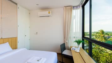 Habitación de hotel con cama, escritorio con silla, aire acondicionado y ventana con vista a follaje.