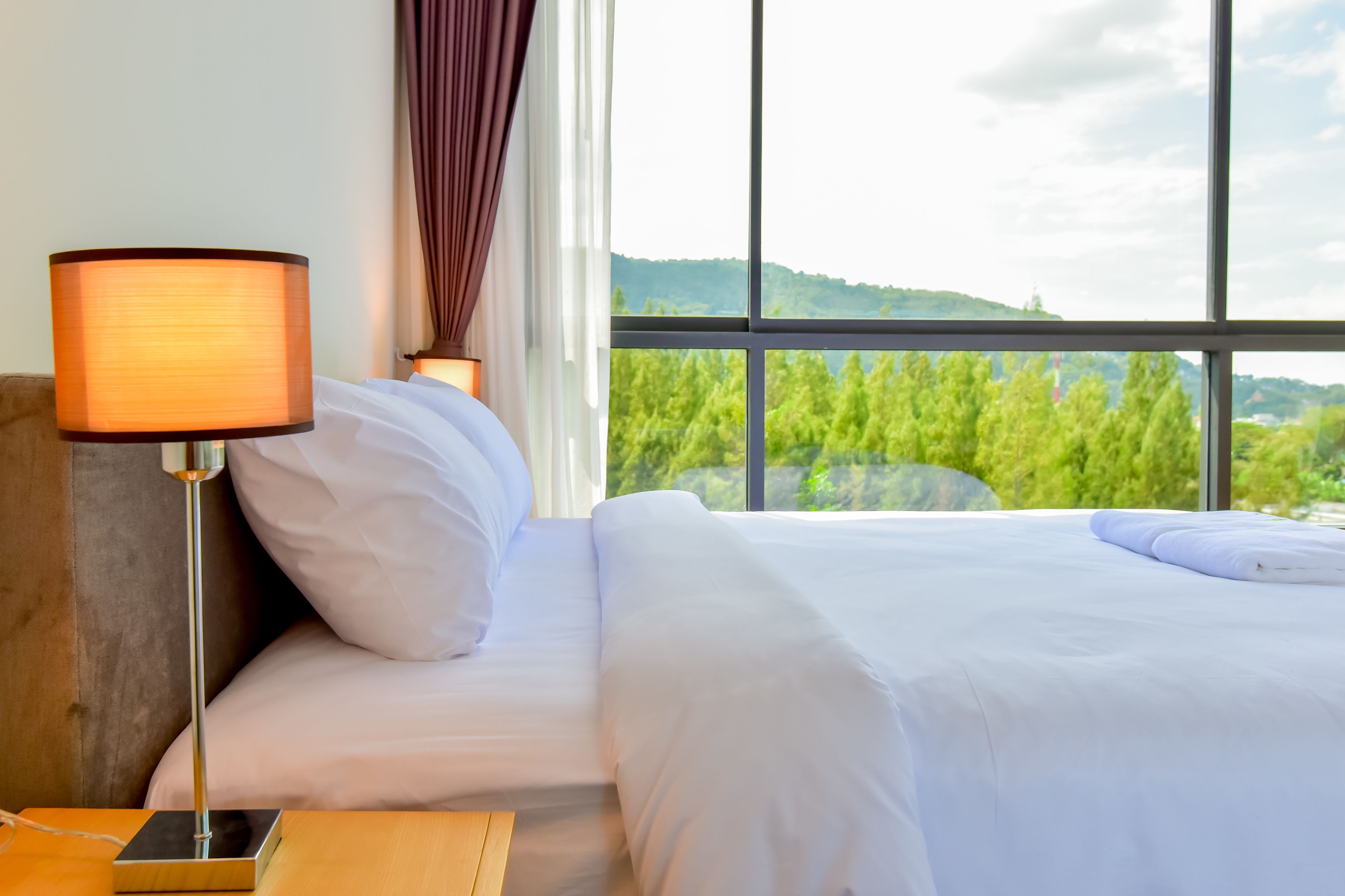 Ein Hotelzimmer mit einem Bett, einer Nachttischlampe, einem Holfnachtkästchen und Blick auf grüne Bäume und Hügel durch große Fenster.
