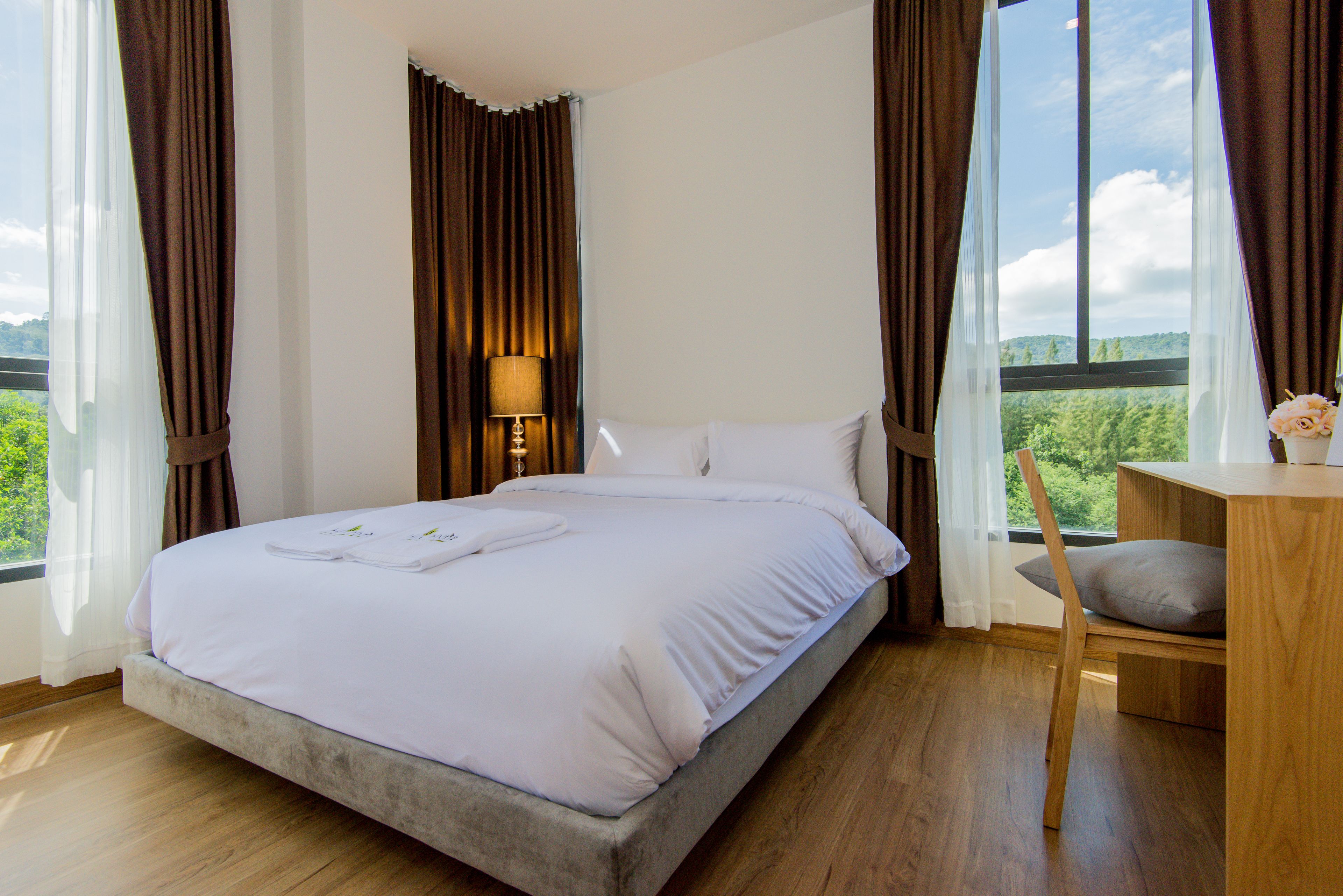 A bedroom with a bed, a bedside table with a lamp, a chair, and a view of greenery through large windows.