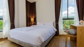A bedroom with a bed, a bedside table with a lamp, a chair, and a view of greenery through large windows.