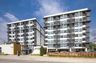 Un edificio residencial moderno de varios pisos con balcones, un estacionamiento y un pequeño local comercial en la entrada.