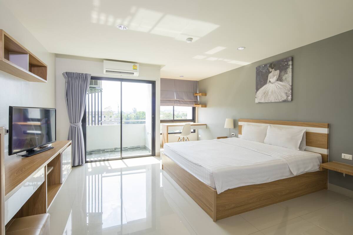 A modern hotel room with a large bed, a TV, a desk, and a balcony with curtains.