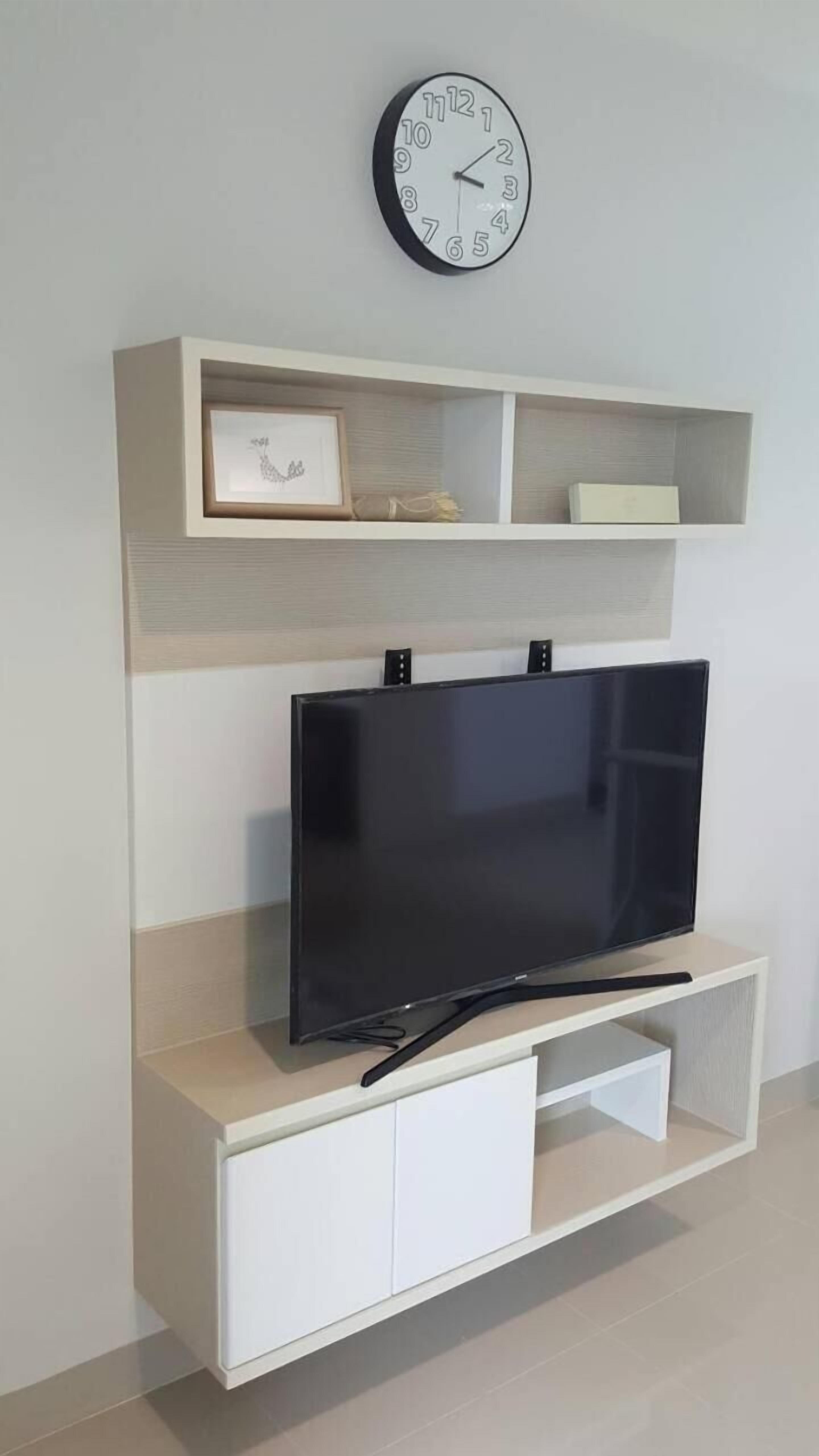 A modern living room with a flat-screen TV on a white entertainment unit, a wall-mounted clock above it, and a framed picture on the shelf.