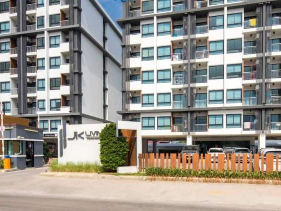En modern flervåningsbyggnad med bostäder, balkonger, en tydligt markerad entré och en logotyp för organisationen ”Amazing Thailand AddHealth & Safety Administration”.