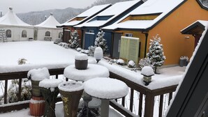 Un espacio exterior cubierto de nieve con una terraza de madera, plantas en macetas y un edificio con techo inclinado.