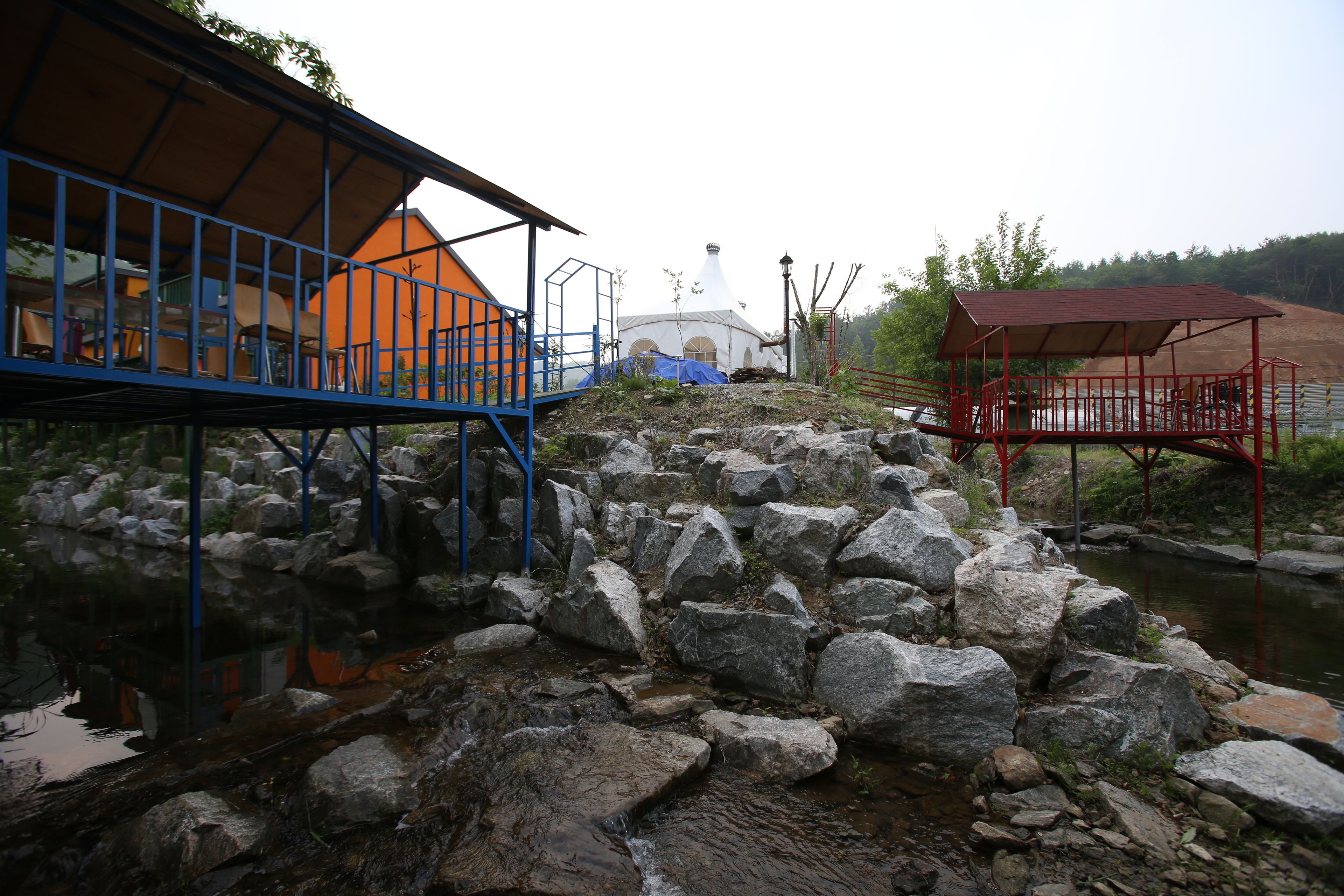 Un espacio al aire libre rústico con un puente de madera, una estructura en forma de cúpula blanca y un pabellón rojo.