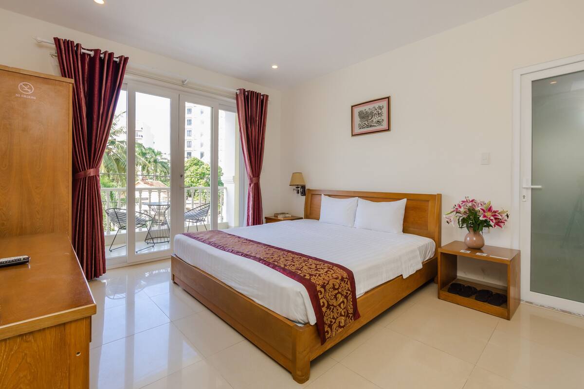 A hotel room with a large bed, a wooden desk, a nightstand with a lamp, a vase with flowers, and a sliding glass door to the balcony.