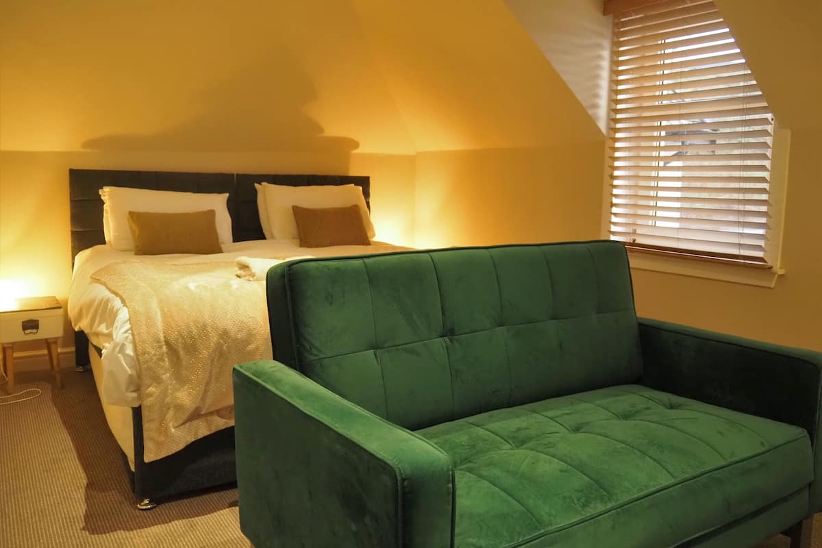 A room with a bed, a green armchair, a nightstand, and a window with blinds.