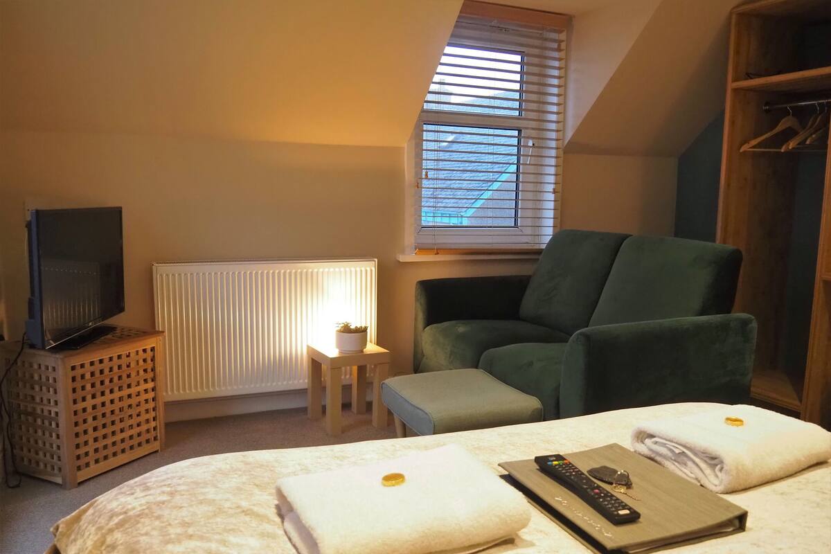 A hotel room with a bed, a green armchair, a small table with a plant, a television on a wooden stand, and a window with blinds.