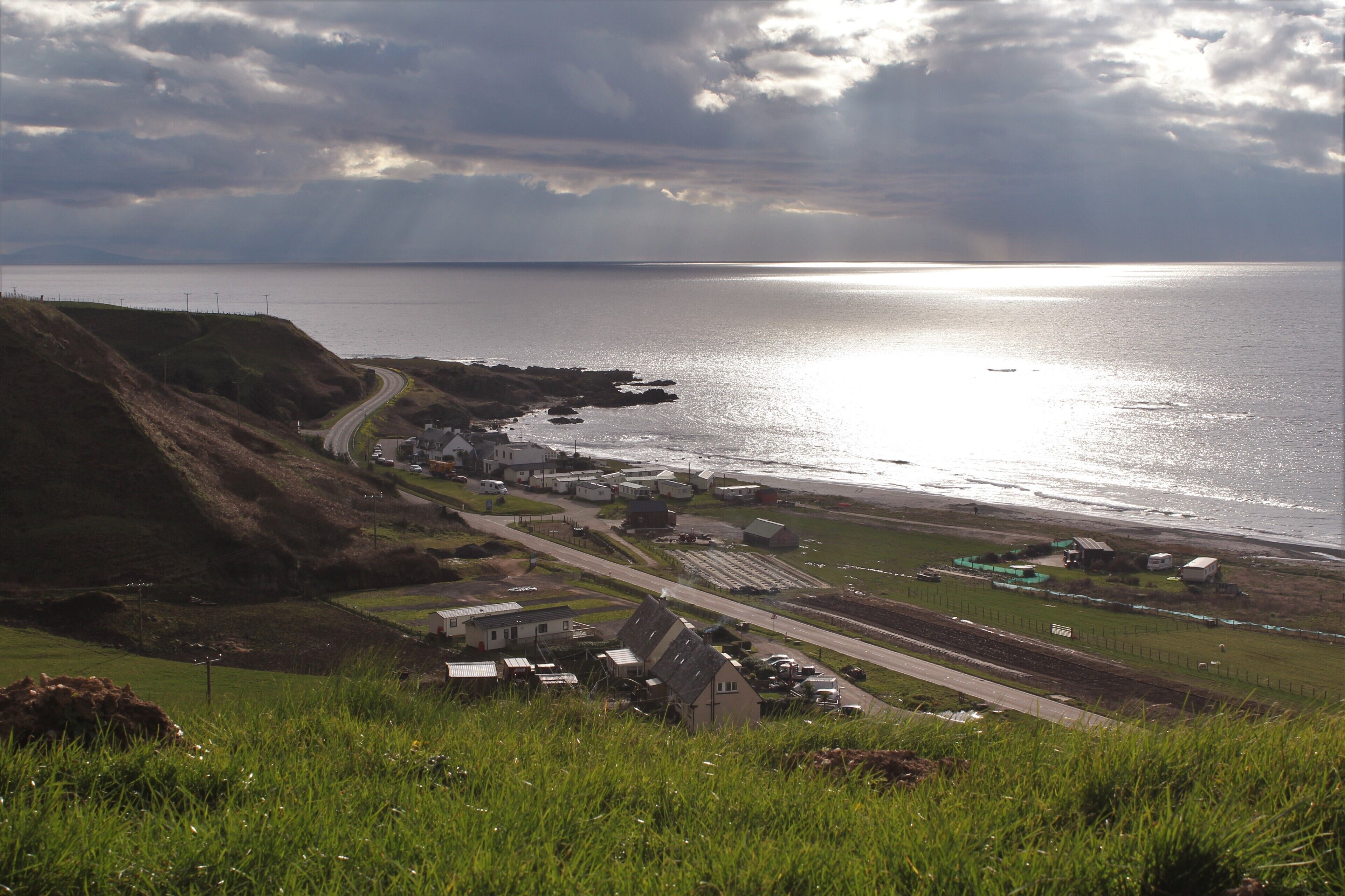 A coastal town with a winding road, houses, and a view of the sea under a cloudy sky.