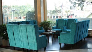Un área de descanso con sillas tapizadas en color turquesa y una mesa de centro, ventanas amplias con vistas a la ciudad y plantas en macetas.
