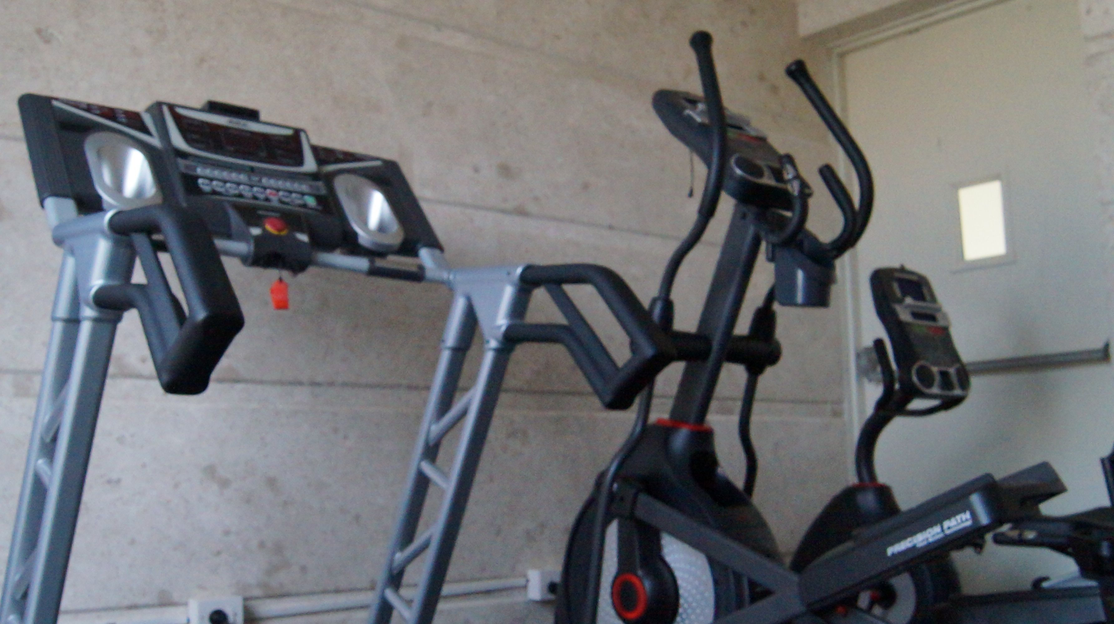 Una bicicleta estática y una cinta de correr en una habitación con una pared lisa y una puerta.