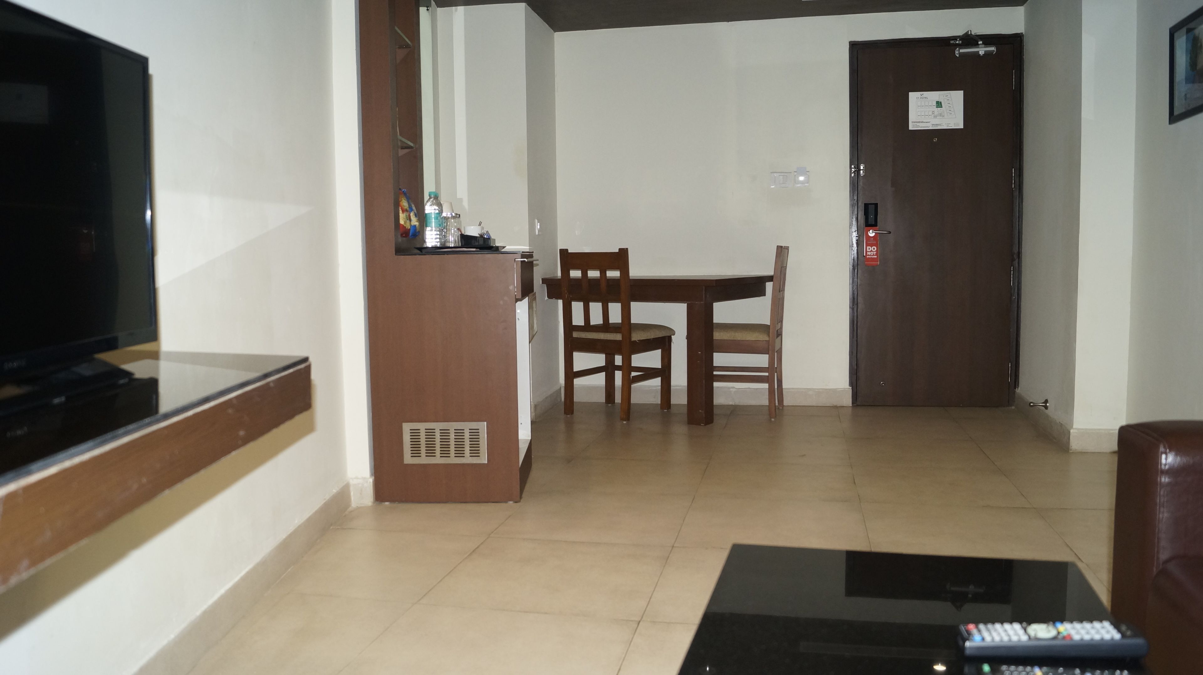 A living room with a flat-screen TV, a wooden table with two chairs, a coffee table, and a door with a 'Do Not Enter' sign.