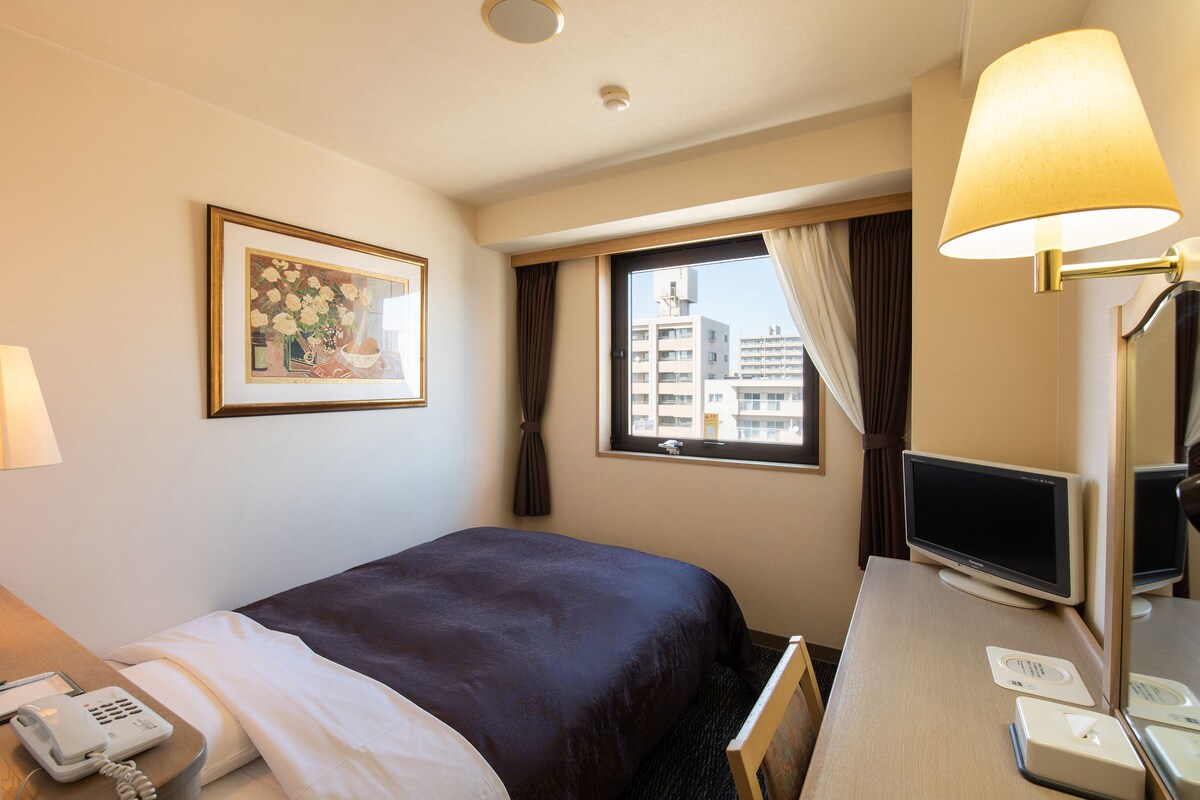 Una habitació d'hotel amb un llit, un escriptori amb un ordinador, un telèfon, un quadre amb marc a la paret i una finestra amb cortines.