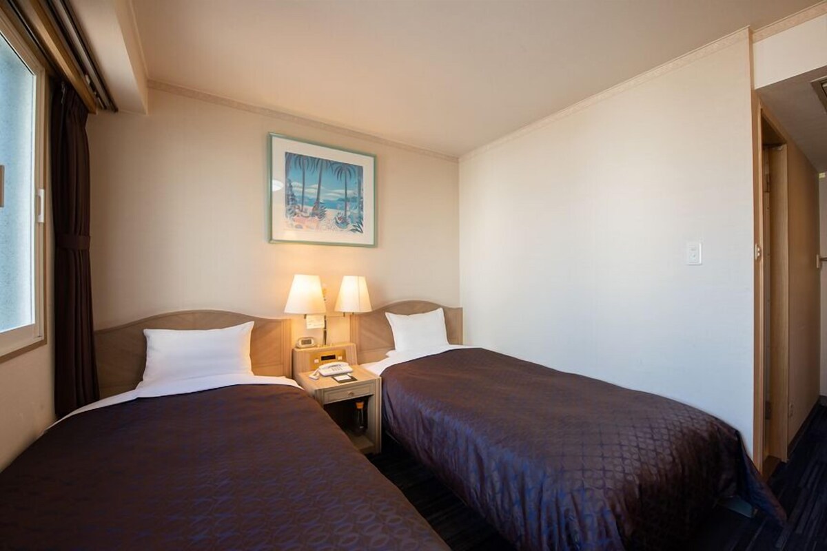 A hotel room with two beds, a nightstand with lamps, a framed picture on the wall, and a window with curtains.