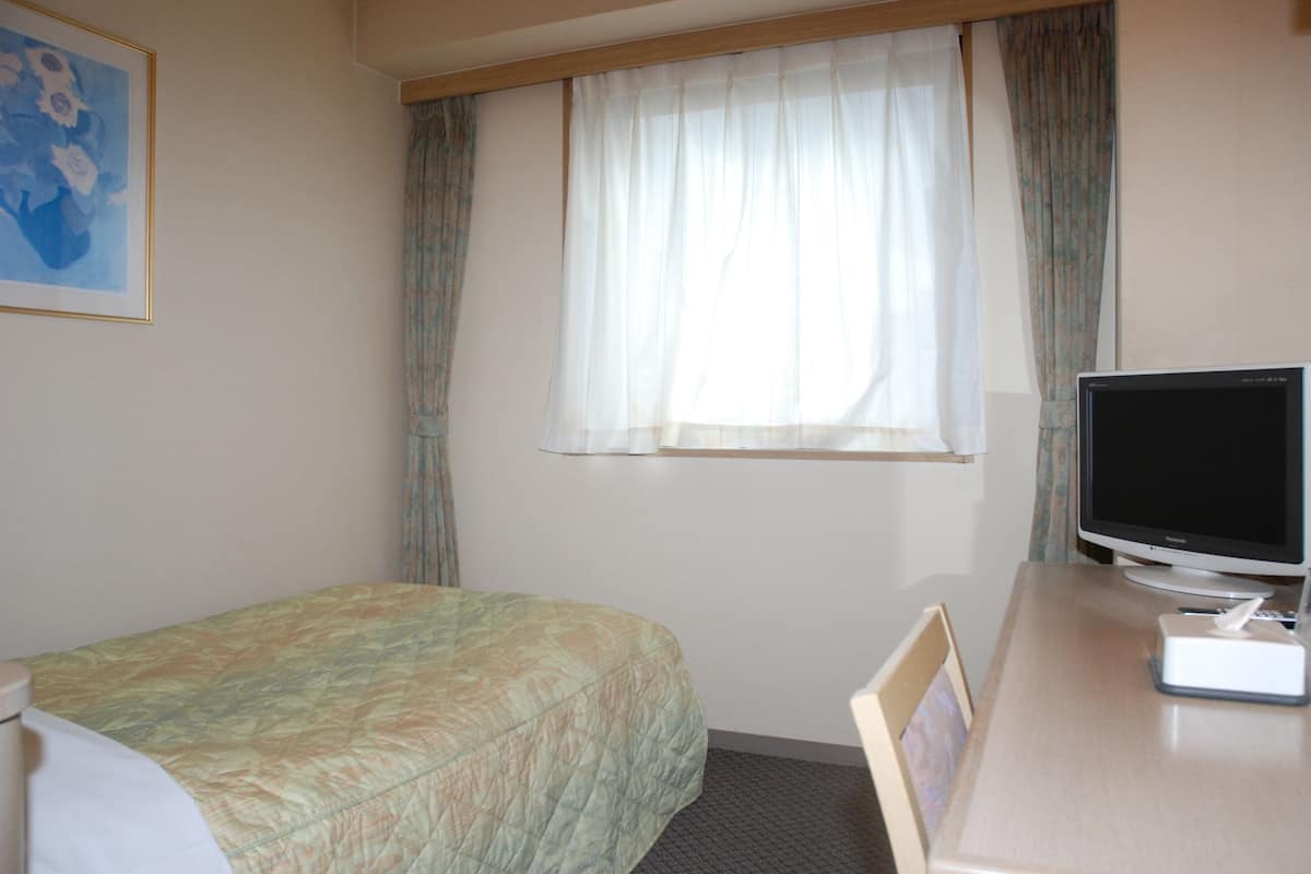 A hotel room with a bed, a desk with a computer monitor, and a window with curtains.