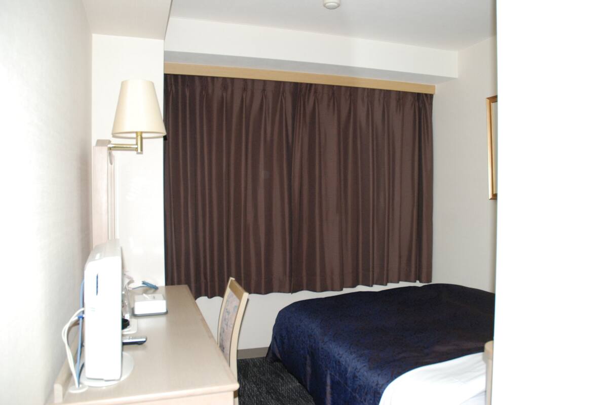 A hotel room with a bed, a desk with a lamp, and a window with curtains.