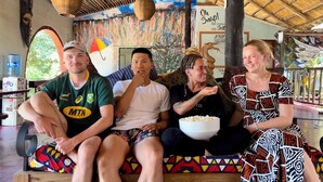 A group of people sitting on a bench in a covered outdoor area with a thatched roof, colorful wall art, and a water dispenser in the background. - Jollyboys Backpackers (Livingstone)