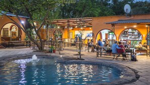 An outdoor dining area with a fountain, tables, and chairs, surrounded by trees and a building with arches. - Jollyboys Backpackers (Livingstone)