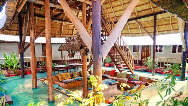 An open-air lounge with wooden pillars, colorful seating, and a thatched roof. - Jollyboys Backpackers (Livingstone)
