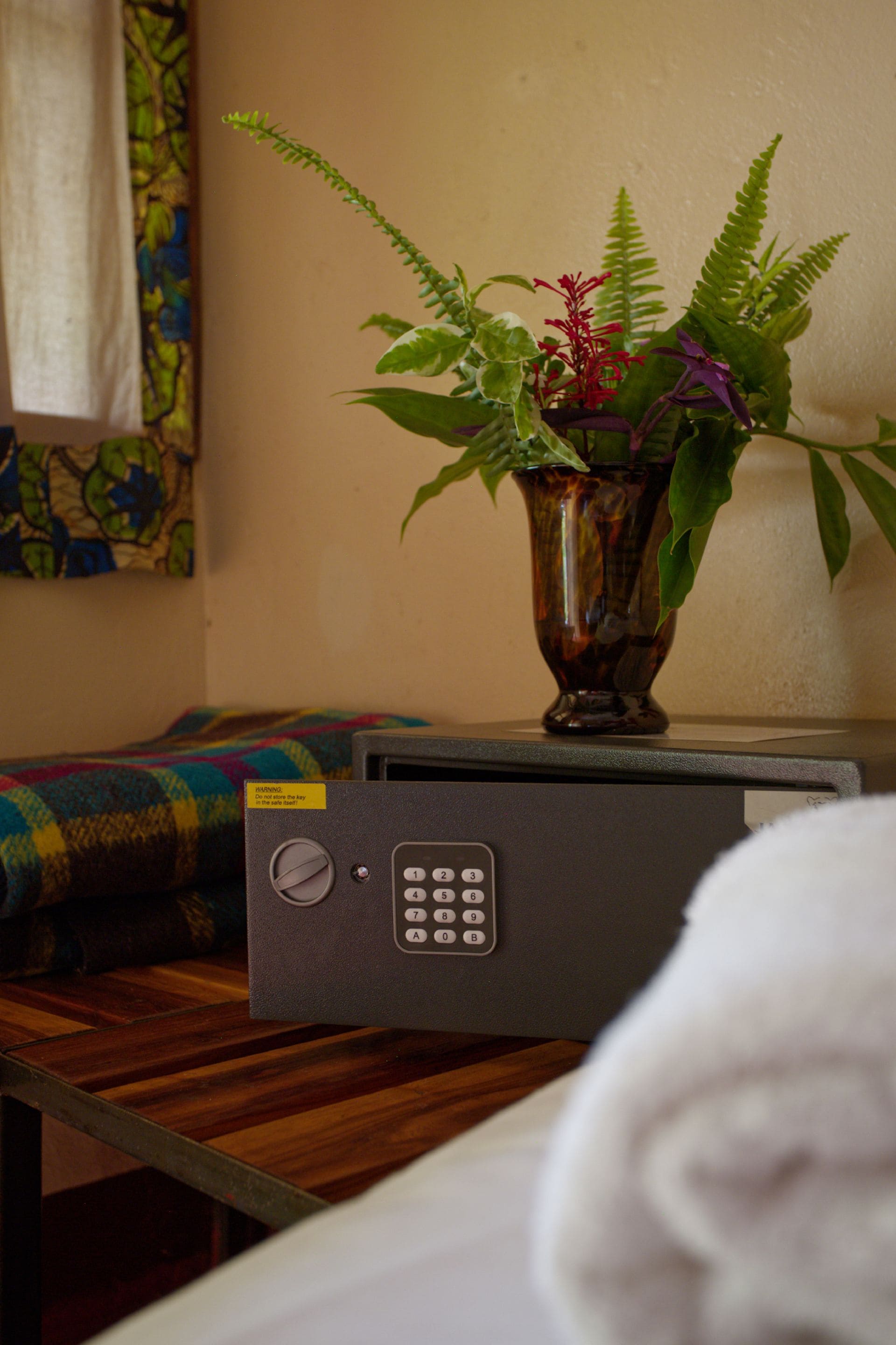 A room with a bed, a small table with a vase of plants, and a safe on the table.