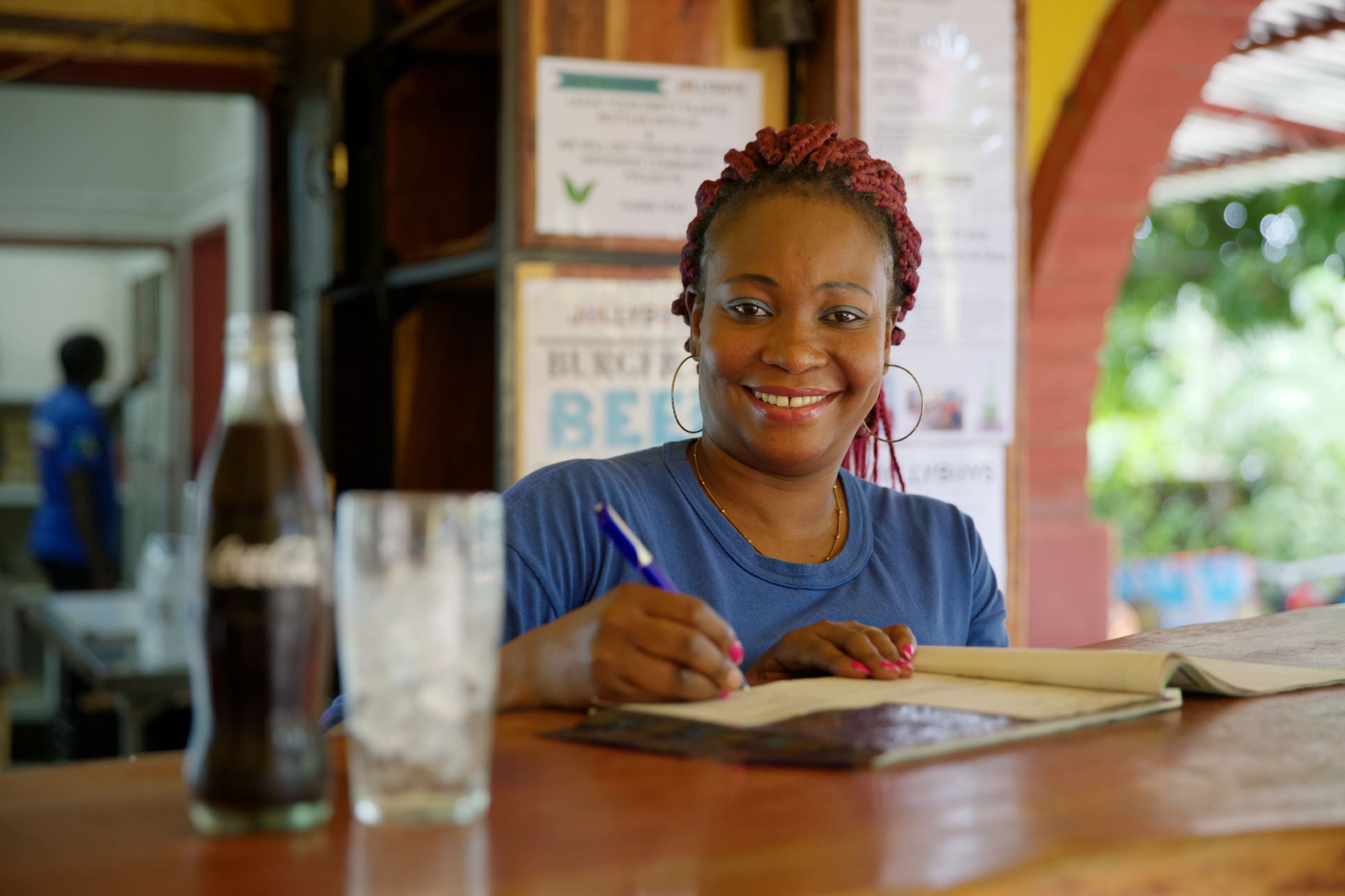 A person sitting at a wooden table with a notebook and pen, a bottle of Coca-Cola, and a glass of water.
