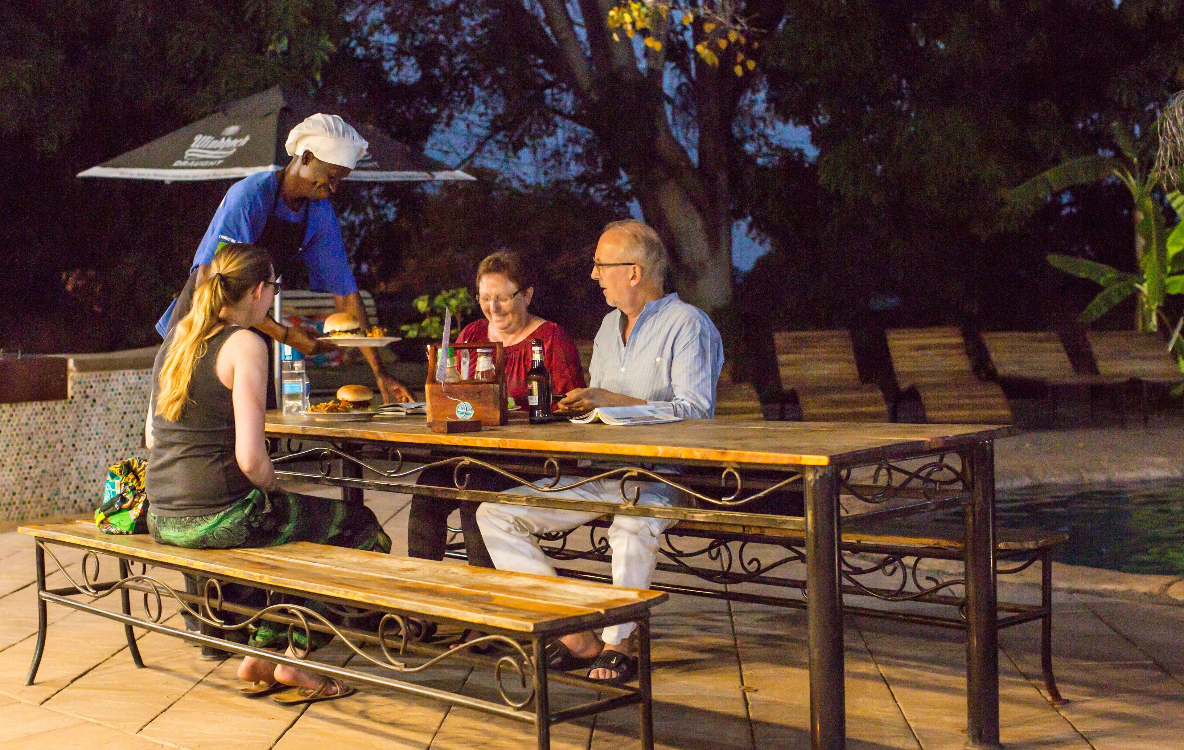 A dining table with benches, a waiter serving food, and a pool in the background.