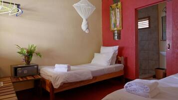 Habitación de hotel con dos camas, una mesita de noche con radio, una planta junto a la cama, una mosquitera y una pared roja con un cuadro.