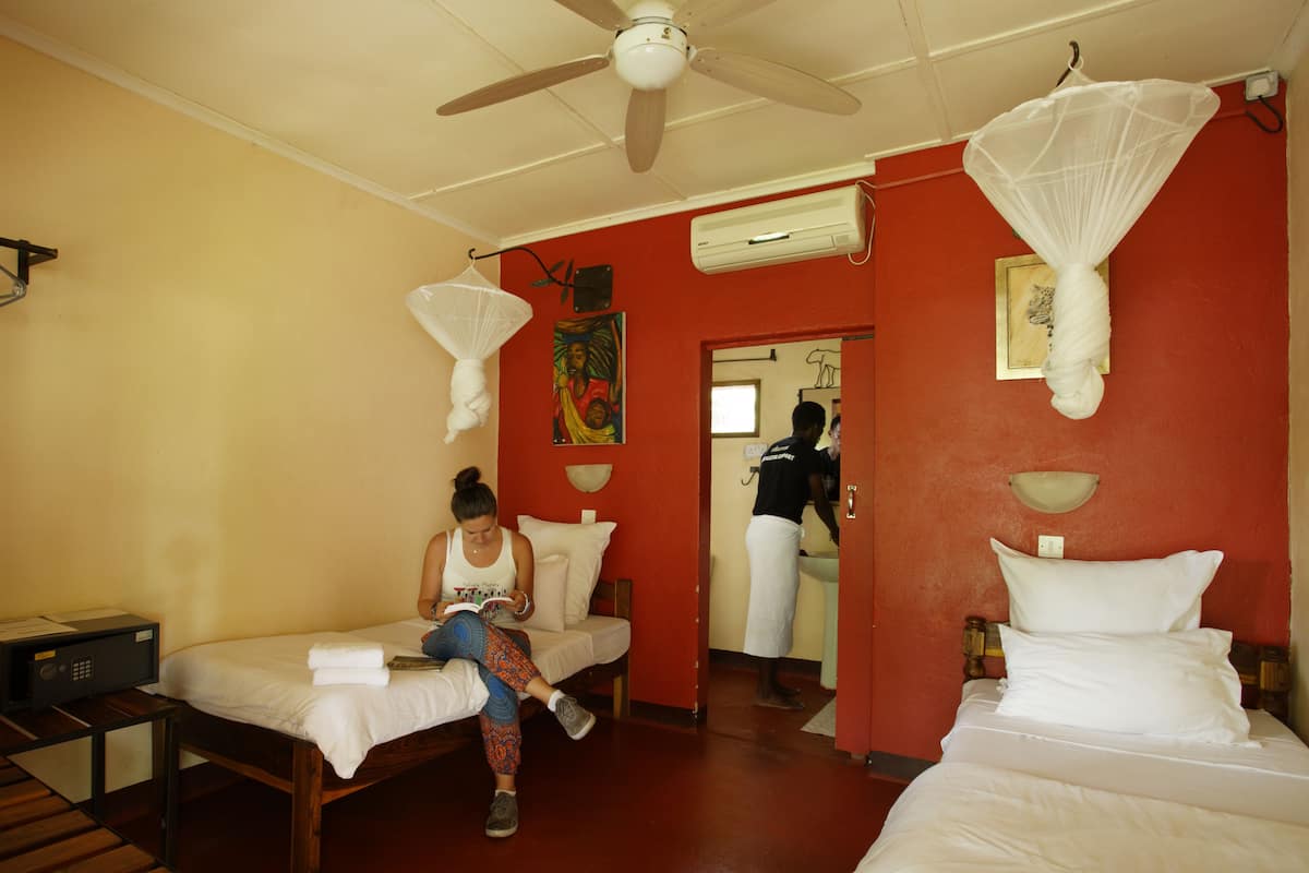 A hotel room with two beds, a ceiling fan, mosquito nets, an air conditioning unit, and a person reading a book.