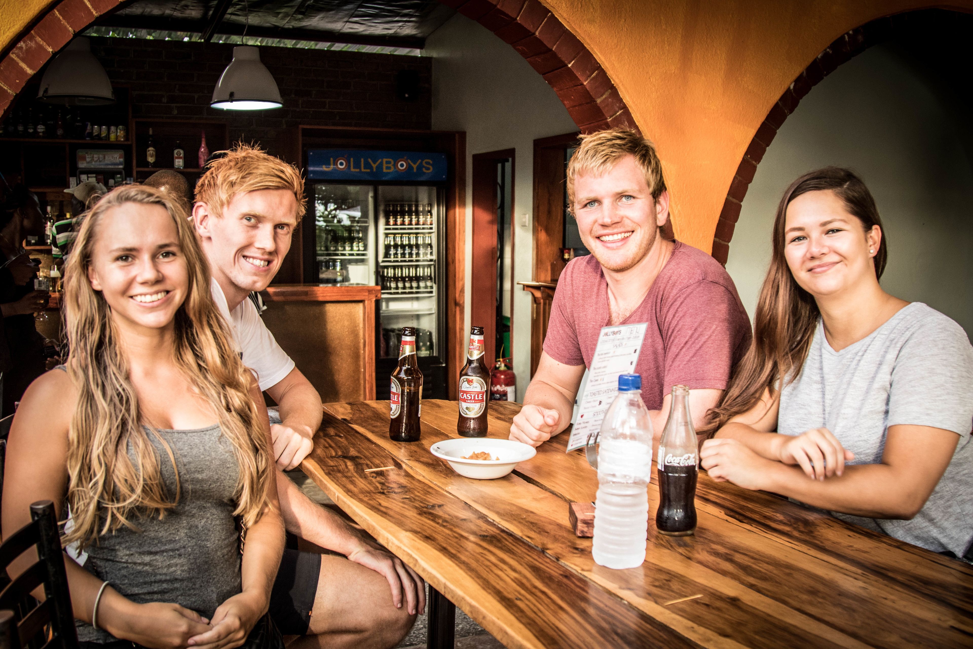 Un grupo de personas sentadas en una mesa de madera en un bar con pared de ladrillo y un letrero que dice "JOLLYBOYS".
