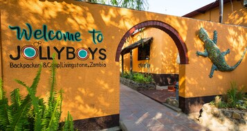 Una señal de bienvenida para Jollyboys Backpackers & Camp en Livingstone, Zambia, con un mural de lagarto y un camino que lleva a la entrada.