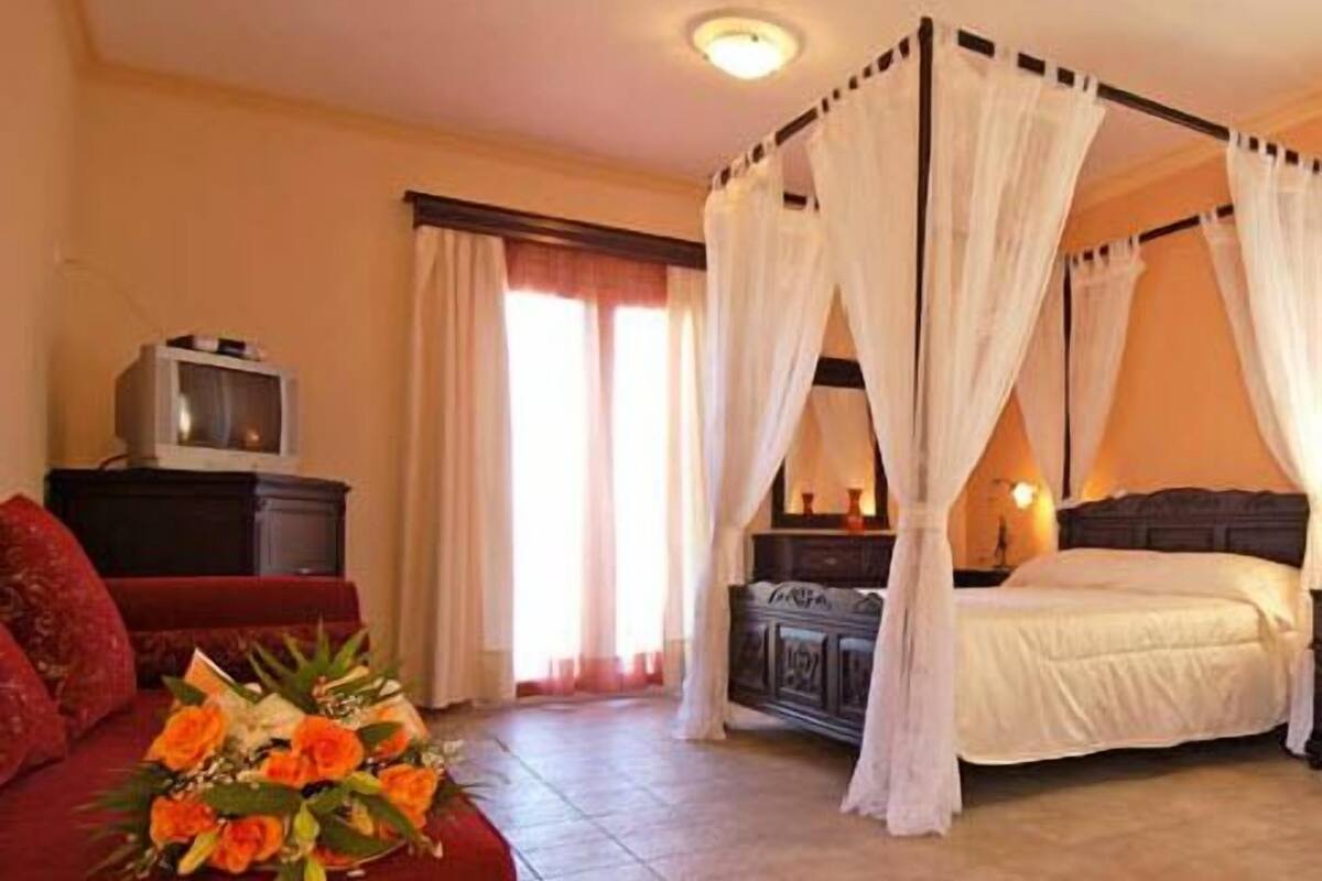 A room with a four-poster bed, a television on a cabinet, and a floral arrangement on a red sofa.