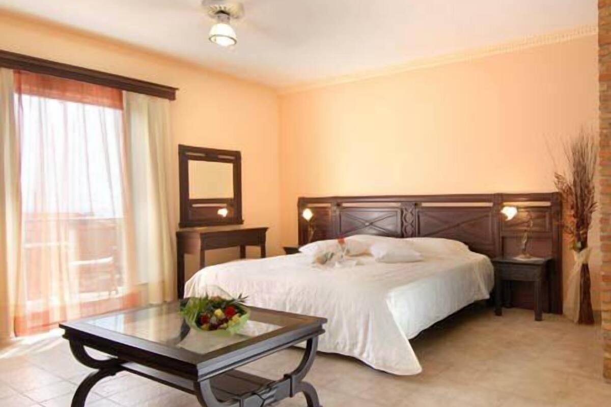A hotel room with a bed, bedside tables, a nightstand, a table with a bouquet, a mirror, and a ceiling fan.